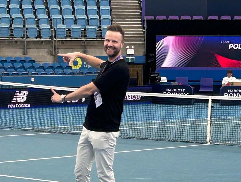 Smiling man standing on the court at United Cup Tennis event pointing at the arena setup