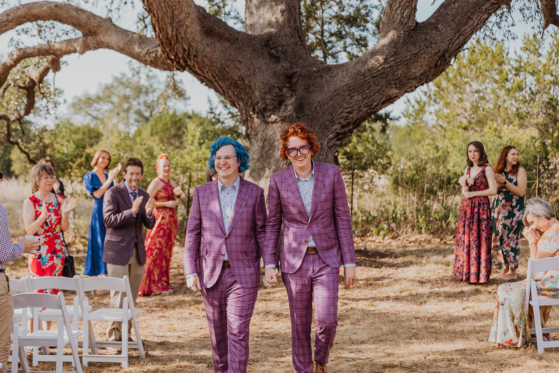 Two grooms in matching suits walk down the aisle at their wedding at Felton Ranch in Dripping Springs, Texas