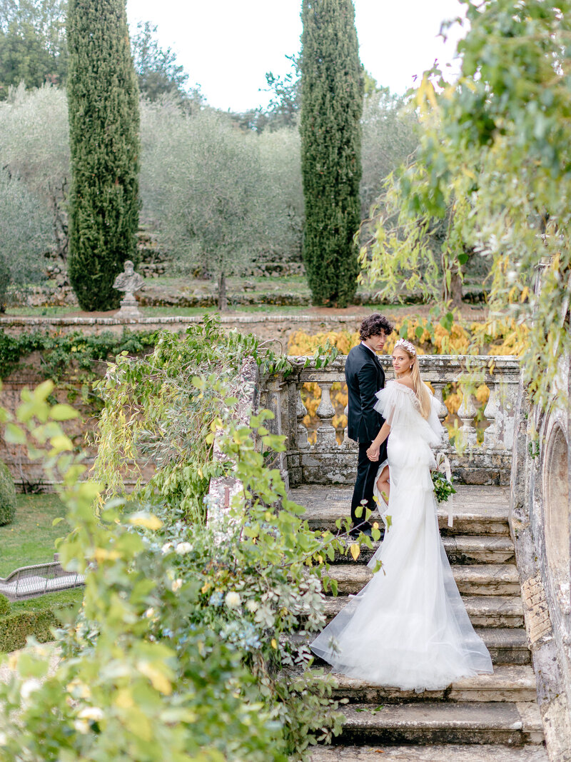 Bride and groom climbing stone villa stairs with cypress trees and olive grove in the background and vines in the foreground