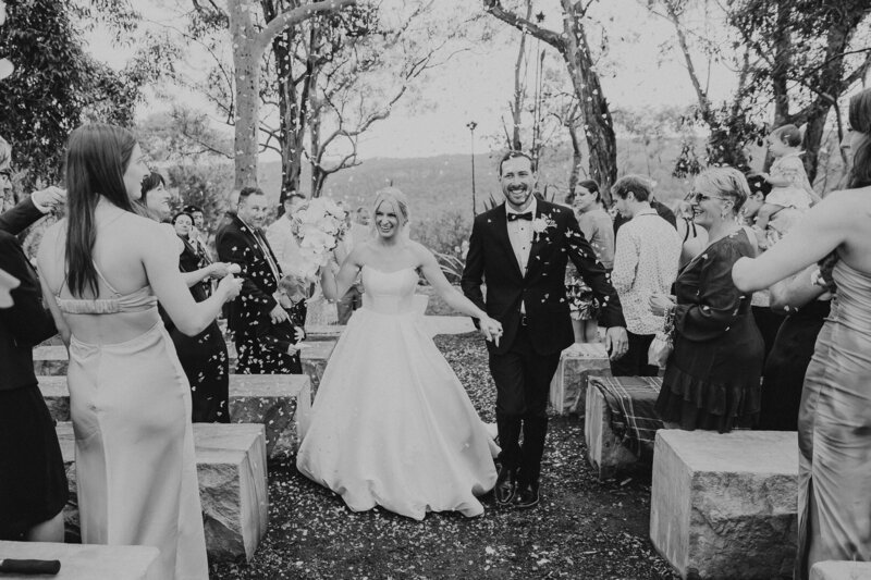 Black and white wedding image of a couple walking up the aisle. They are laughing and smiling while their guests throw confetti over them. Shot at Glenworth Valley, Central Coast Wedding Photographer