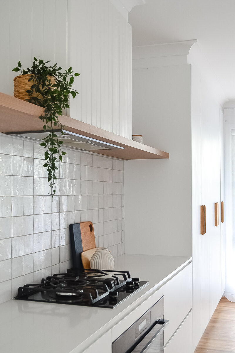 Modern kitchen with a gas stove, white tile backsplash, wooden shelf with a plant, and white cabinets crafted by Bespoke Interior Design Newcastle NSW.