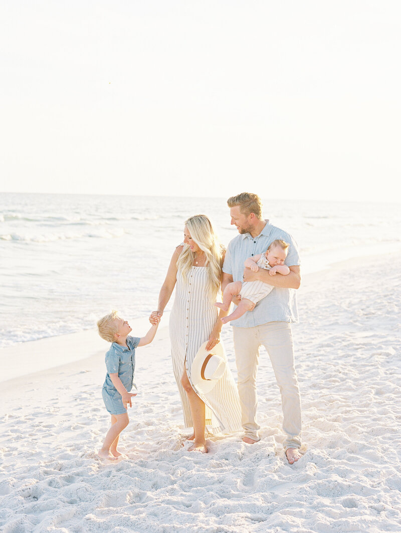 Dad holding baby while mom holds son's hand on the beach with ocean in the background