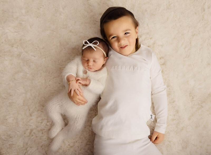 Brooklyn NY newborn photoshoot. Aerial image. Baby boy is smiling at the camera with his baby sister resting on his shoulder. Baby girl is sleeping. Neutral cream palette. Captured by best Brooklyn, NY newborn photographer Chaya Bornstein.
