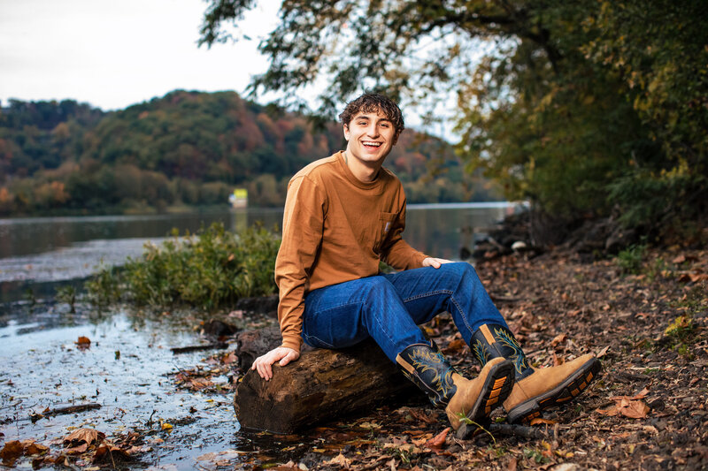 Smiling high school senior boy wearing cowboy boots posing near the river during fall.
