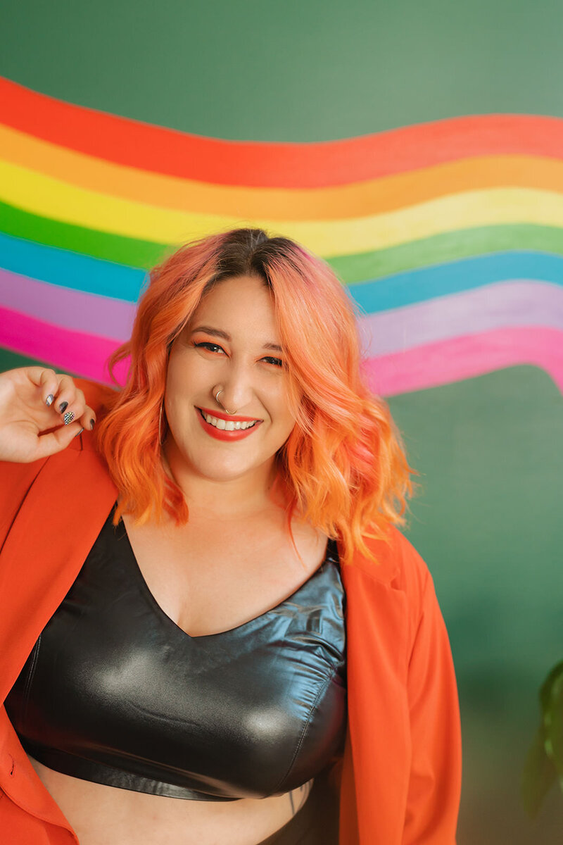 A person with vibrant hair smiles against a rainbow mural