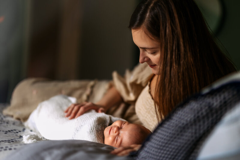 A mom is laying on a bed looking down at a newborn baby wrapped in a white blanket.