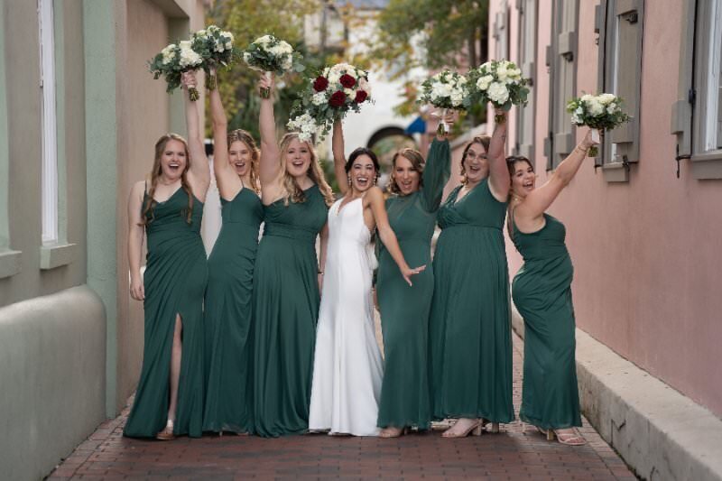 A joyful bride in a white dress celebrates with six bridesmaids wearing matching emerald green gowns, all raising their white and burgundy bouquets in the air while standing on a brick sidewalk in front of colorful historic buildings in downtown St. Augustine