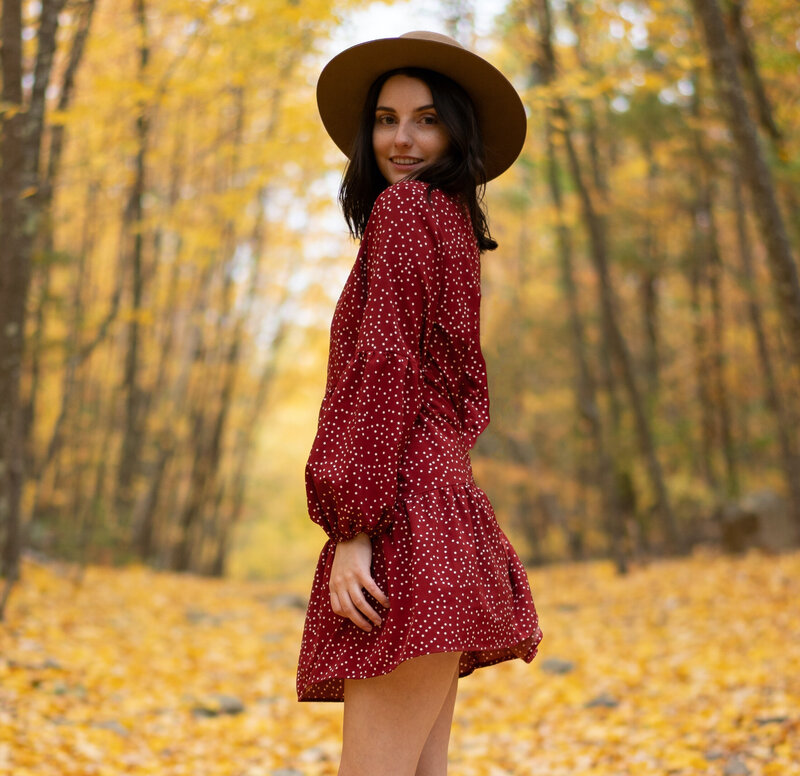 Woman standing in the woods with a red dress wearing a tan hat.