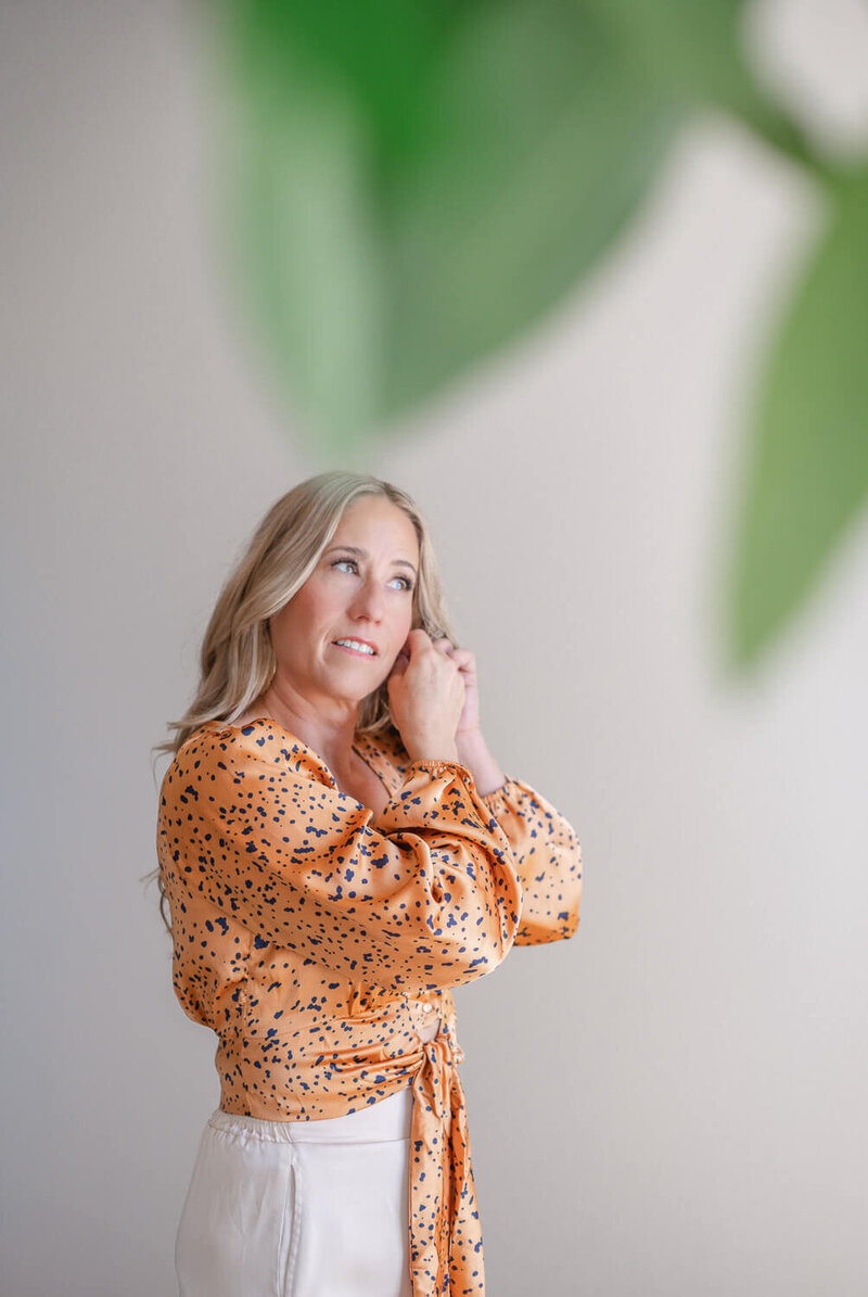Kelowna Brand Photographer, Kristen wearing tan shirt putting on earring against neutral wall. 