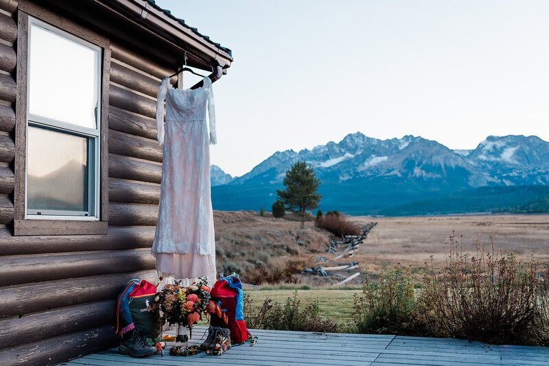 A wedding dress hangs on the side of a well appointed log-sided cabin included in a luxury adventure elopement package at a national park.  a field with a zigzag rail fence is visible in the distance, and behind that a row of jagged topped mountains erupt and fill the skyline. 