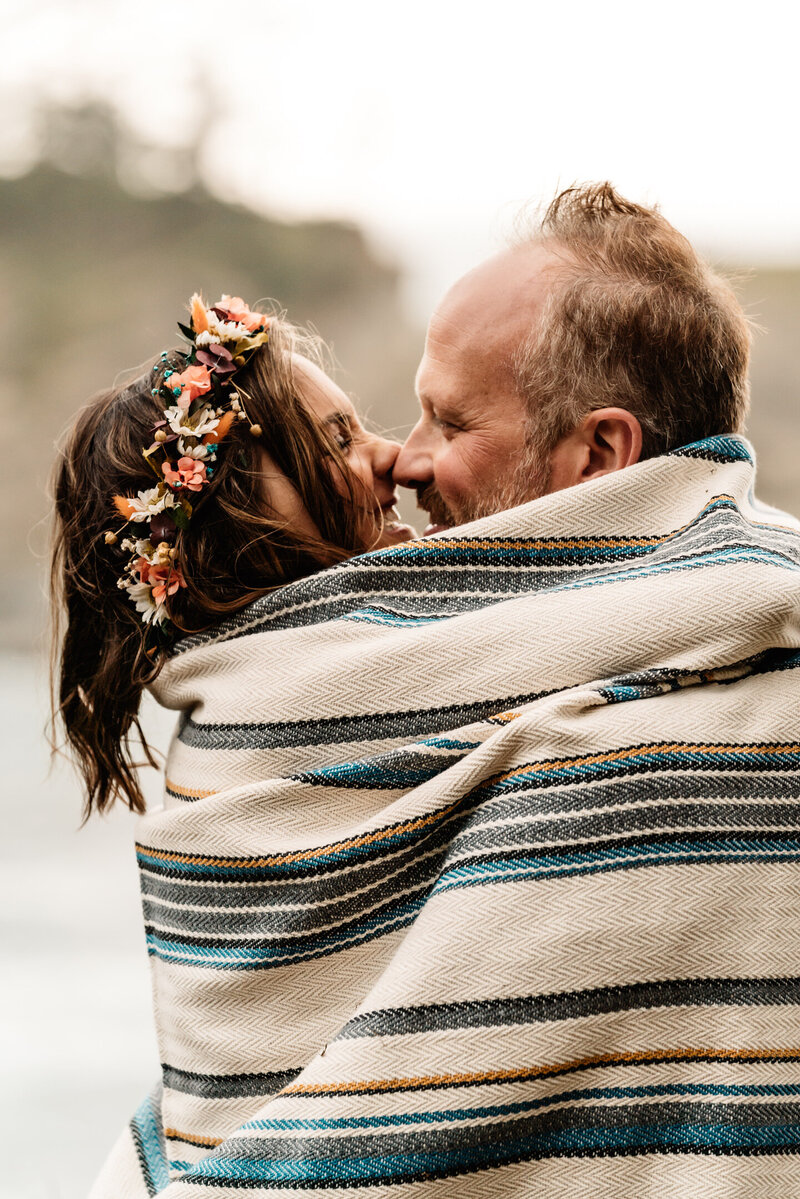 During their Oregon coast elopement, a bride and groom snuggle under a knitted  beach blanket. They rub their noses together and smilee widely at each other.
