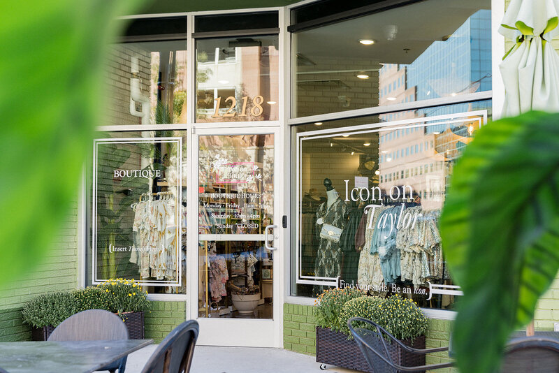 Charming storefront of Icon on Taylor, a boutique in Columbia, SC, with stylish clothing displayed in the window and elegant signage. Greenery and patio seating frame the entrance, creating a welcoming shopping experience. The reflection of city buildings in the glass blends urban and boutique aesthetics. Small business branding photography in Columbia, SC, highlighting fashion retail and local shopping.