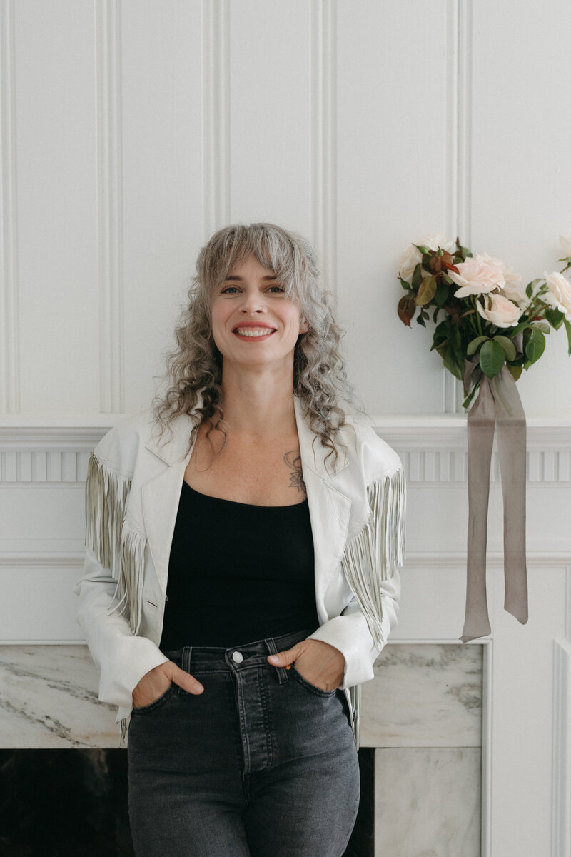 Portrait of a smiling florist with curly gray hair wearing a white fringe jacket and black top, standing in a bright modern studio beside a soft blush rose floral arrangement with flowing ribbon. The image captures a warm, welcoming brand aesthetic for a creative wedding florist.