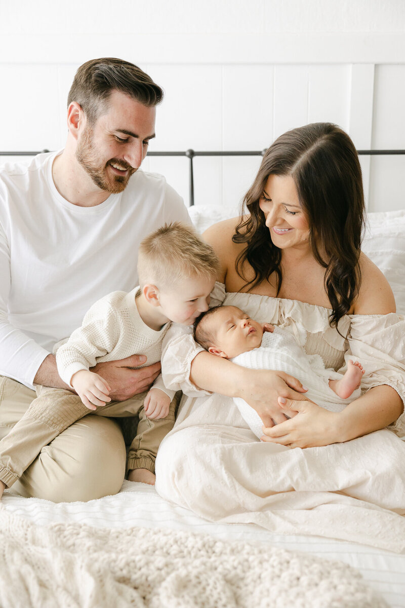 family dressed in cream sits on bed, parents look down smiling as big brother kisses his baby brother on the top of his head, Indianapolis family photographer