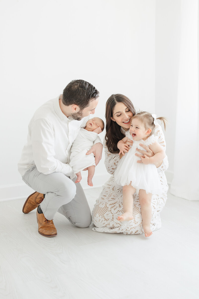 Mom and dad kneel on the floor and smile with their giggly toddler daughter and sleeping newborn during newborn portrait session