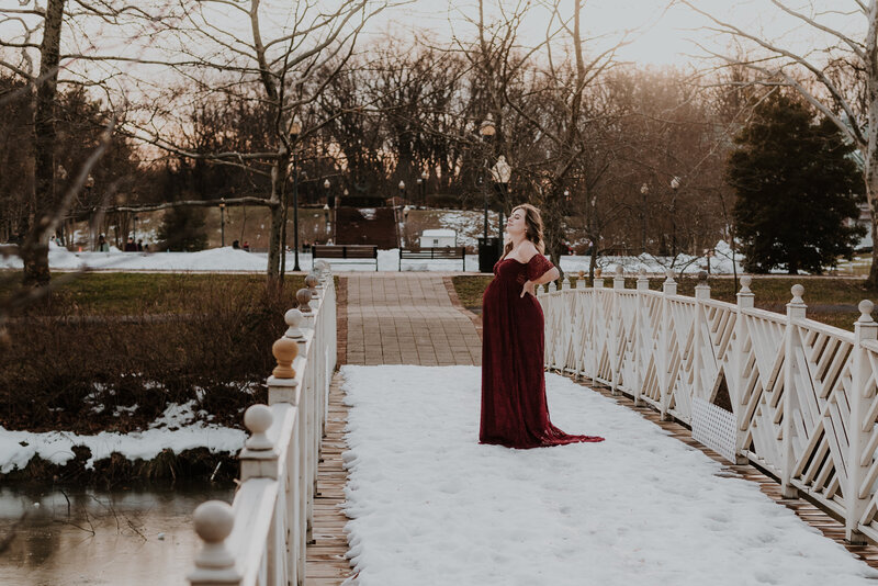 Pregnant mom wearing a red dress standing on a snowy bridge in a garden looking at the sky in Annapolis Maryland.