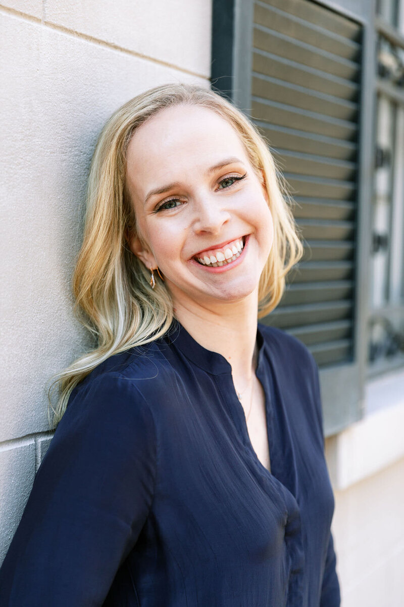 Digital Grace Design owner and Showit web designer, Sarah Blodgett, wears navy blouse and smiles while leaning against a wall outdoors