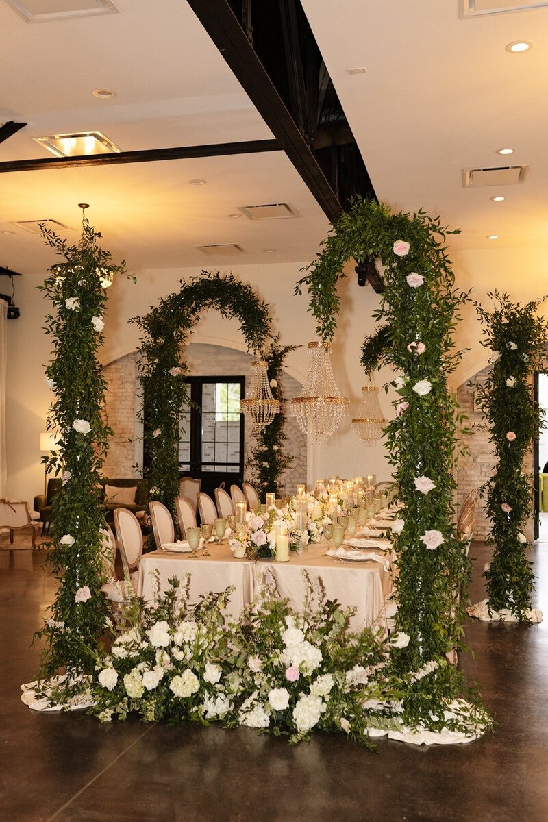 An indoor wedding reception with a long table decorated with candles and floral centerpieces is set beneath green archways with white flowers at grbic events, a wedding venue in st. louis 