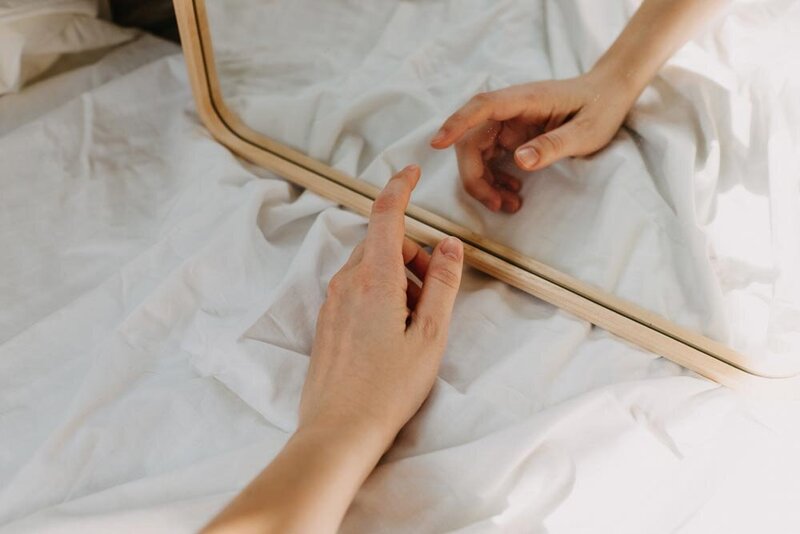 A hand reaches towards its reflection in a wooden-framed mirror, resting on a white, crumpled sheet. The scene conveys introspection and calm.