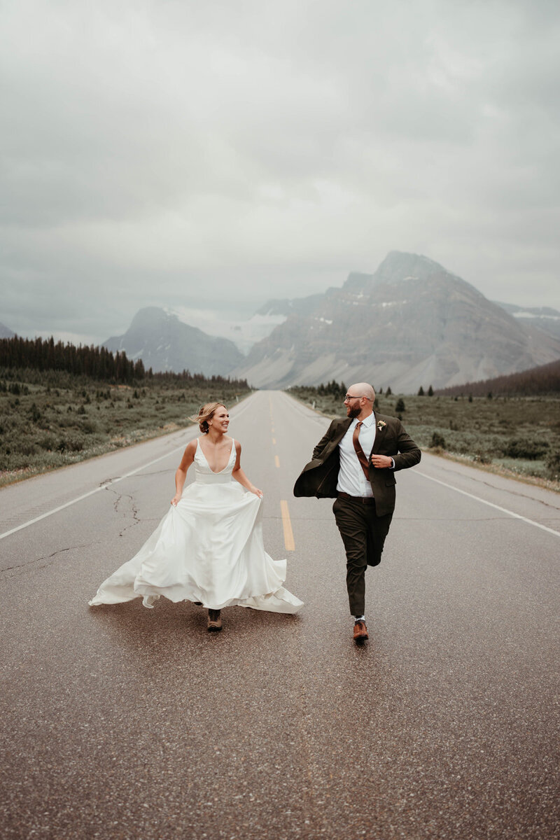 Bride and groom running hand-in-hand down a scenic mountain road in the Canadian Rockies, captured after their wedding ceremony.