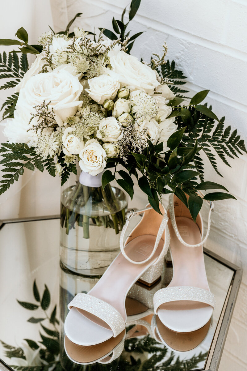 The brides bouquet of white roses and greenery sat beside her white pearl detailed shoes on a mirror end table