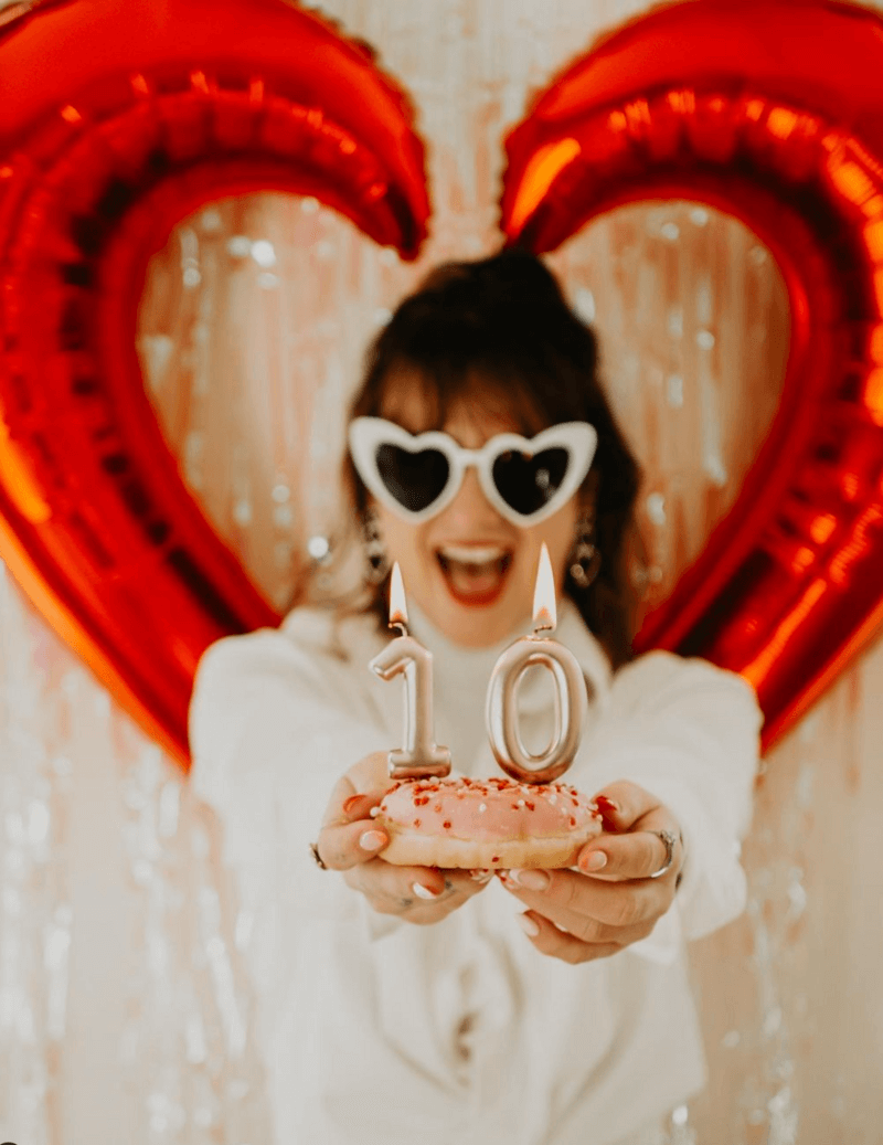 Laura tenant un donut avec des bougies "10 ans", lunettes en cœur. Ballon métallisé rouge en arrière plan en forme de coeur.