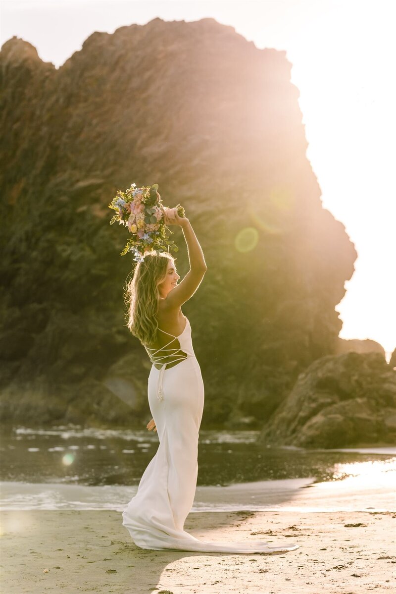 As the sun sets on her Oregon coast elopement, a bride twirls in its rays, her bouquet raised above her head, and a lens flare bouncing in the frame