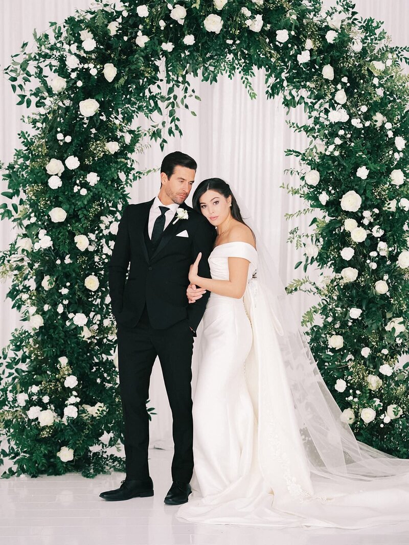 Elegant wedding couple in front of a lush green and white floral arch backdrop, showcasing beautiful ceremony design by a Virginia wedding florist.