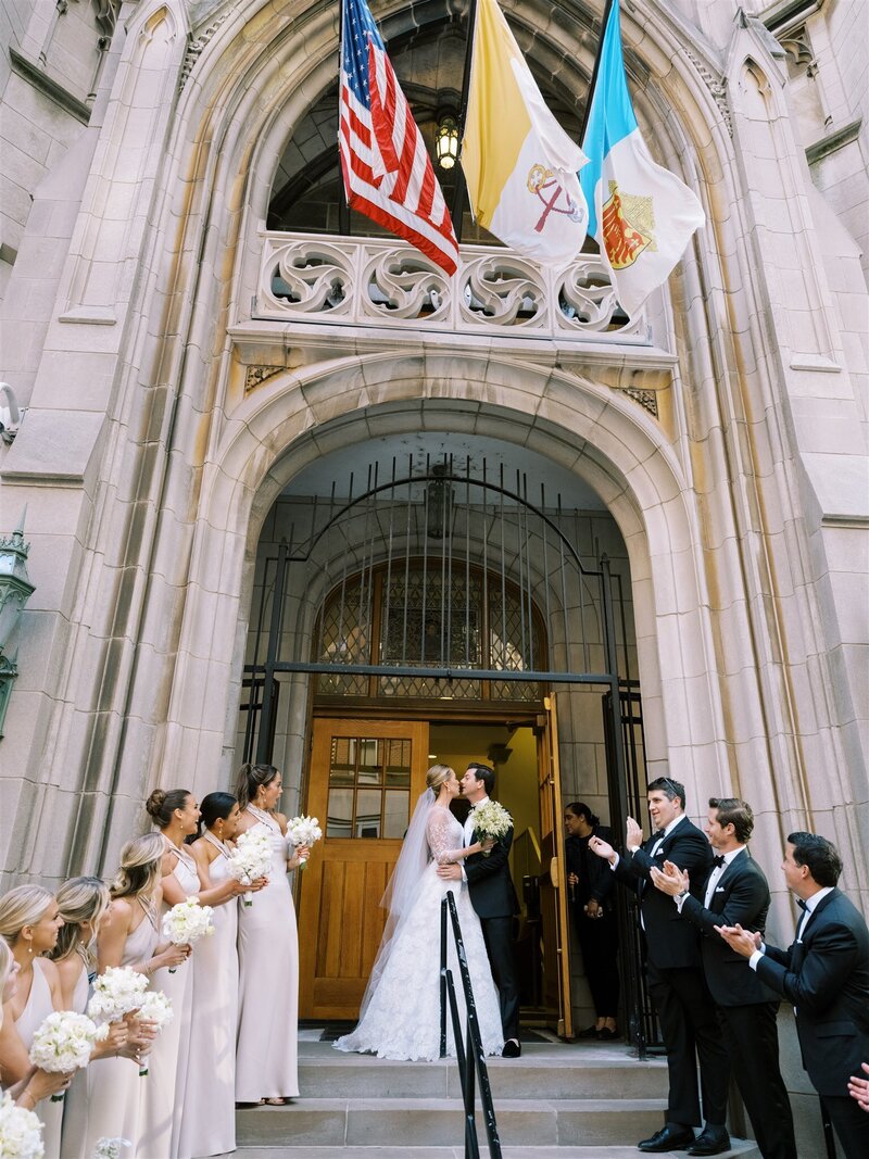 Wedding guests celebrating as a newlywed couple exits the church under an arch of hands.