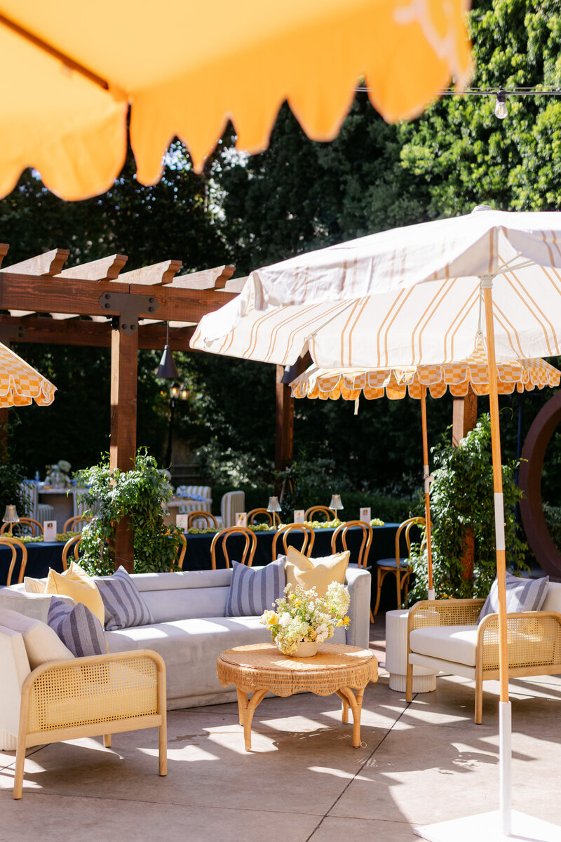 Outdoor seating area with our whale velvet sofa, cream cane accent chairs, and woven scalloped coffee table under our golden check and golden scalloped umbrellas. Background includes a wooden pergola, dining tables, and lush greenery.