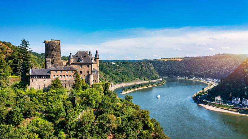 A hilltop medieval castle surrounded by dense green forest overlooking a wide, winding river under a bright blue sky.
