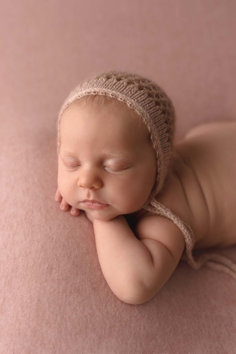 Newborn Baby Girl Laying on Tummy with Head on Hands Posed on a Pink Blanket Wearing a Pink Bonnet