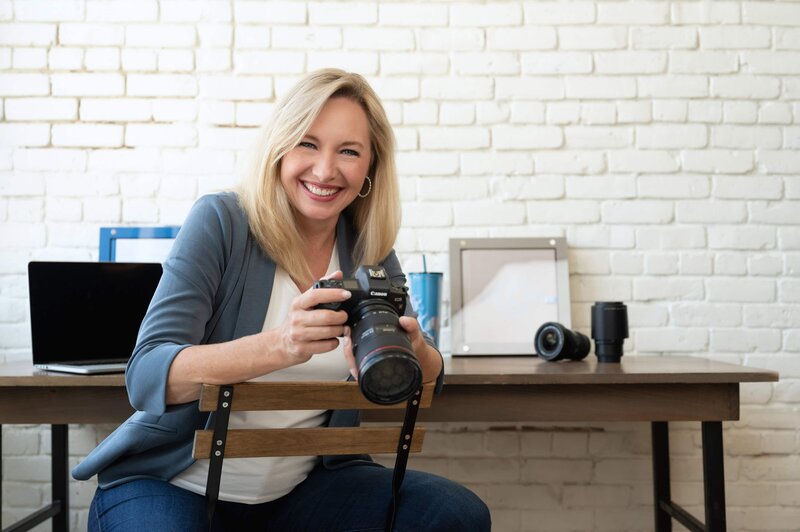 Jen Denton headshot looking at the camera sitting down with her camera in her hand wearing jeans and white jacket