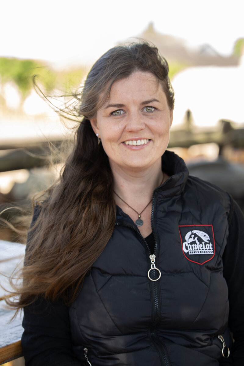 Marise Daimond, instructor at Camelot Horse Riding School, smiling while wearing a Camelot-branded vest beside a horse.