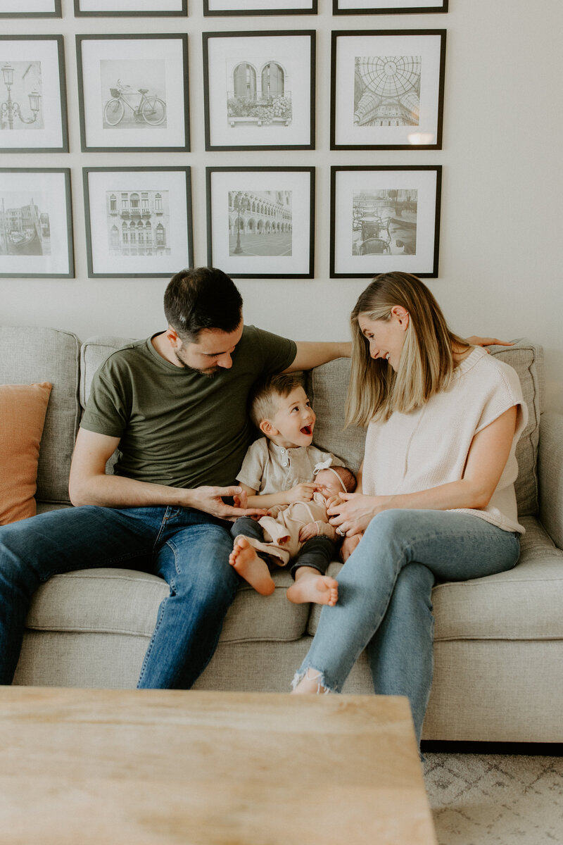 Family sitting on couch with toddler son holding new baby sister during St. Louis newborn photos