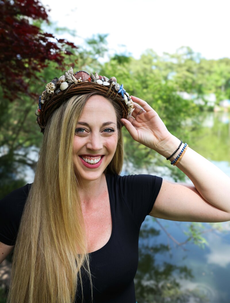 Smiling woman wearing a seashell crown and black top, standing outdoors near a pond with lush green trees and red maple leaves in the background.