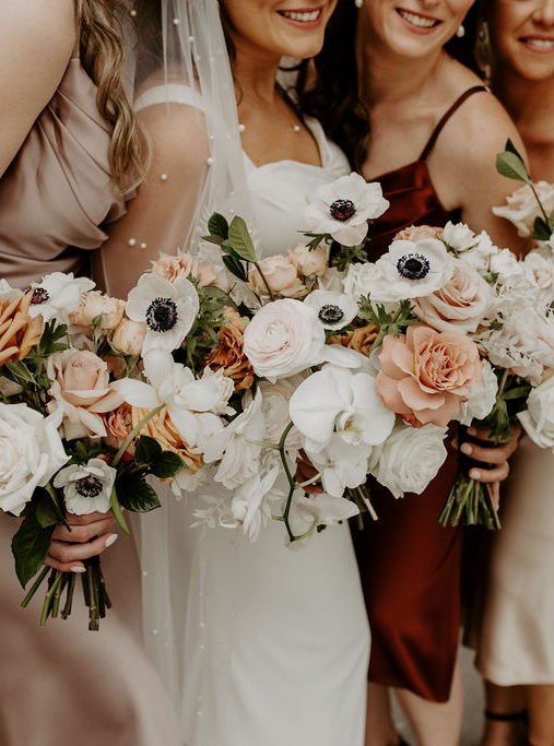 Close-up of a bride and three bridesmaids in earth tones, smiling and holding white, terracotta, black, and blush modern bouquets, capturing the essence of stylish weddings in Ottawa.