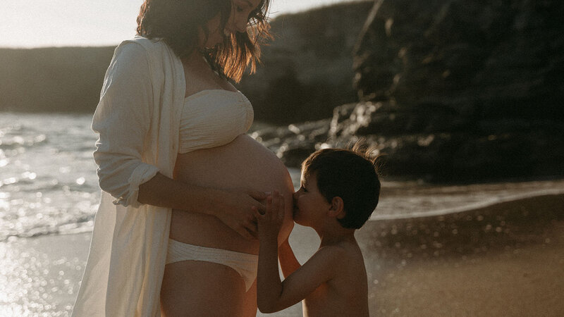 Petit garçon de profil embrassant le ventre rebondi de sa maman à la plage. Séance photo grossesse par Kamescop, vidéaste et photographe en bretagne.
