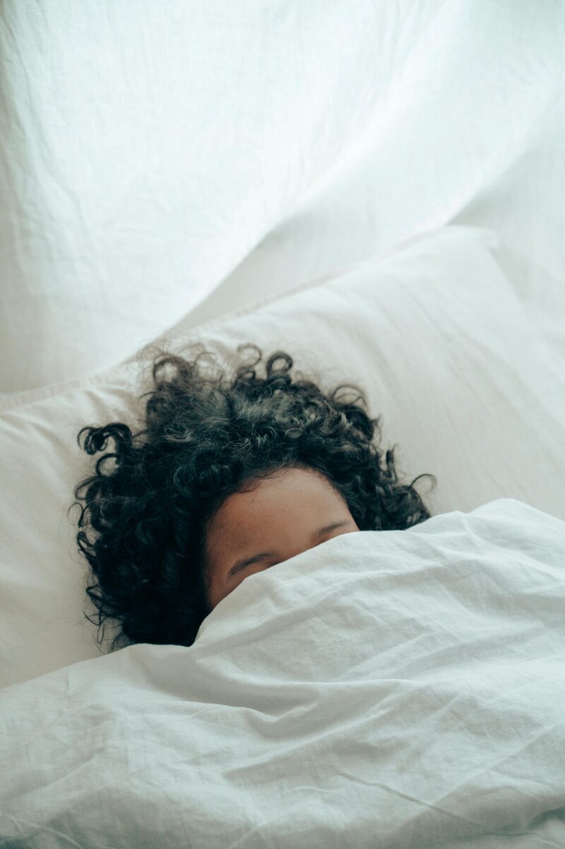 Une femme couchée dans son lit avec les cheveux ébouriffés sur le coussin. Elle est caché par sa couette blanche qui couvre son visage jusqu'aux sourcils. 