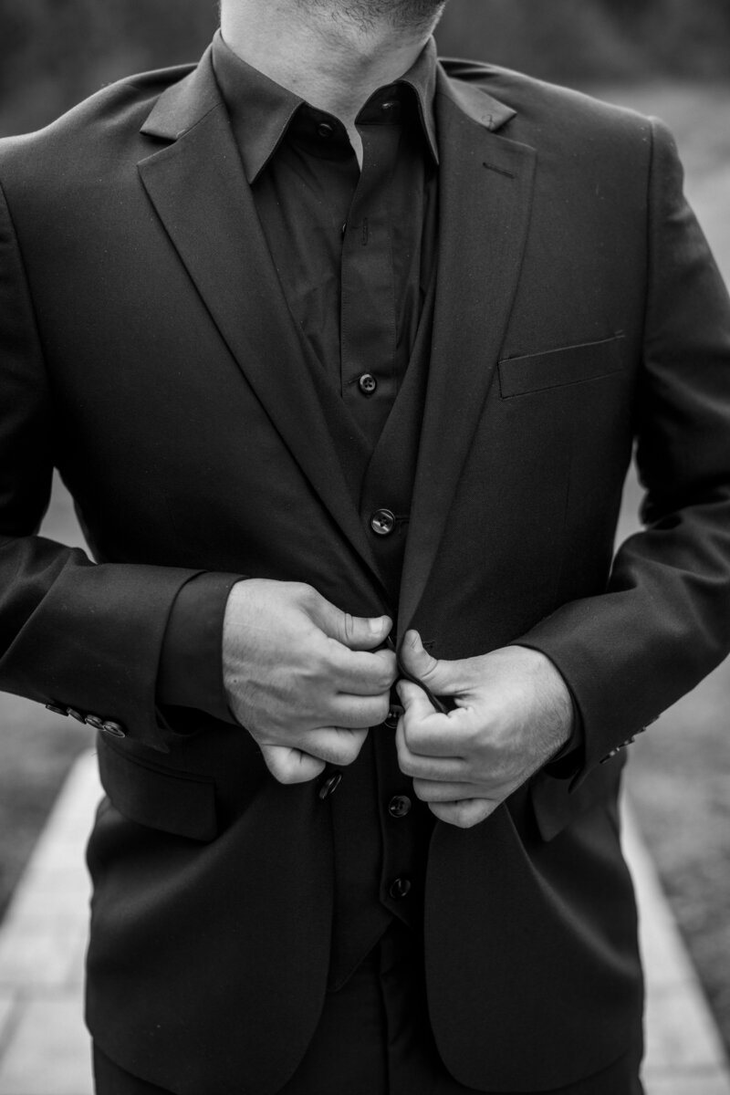 Close-up of groom’s hands buttoning his black suit jacket, a timeless preparation moment before the ceremony
