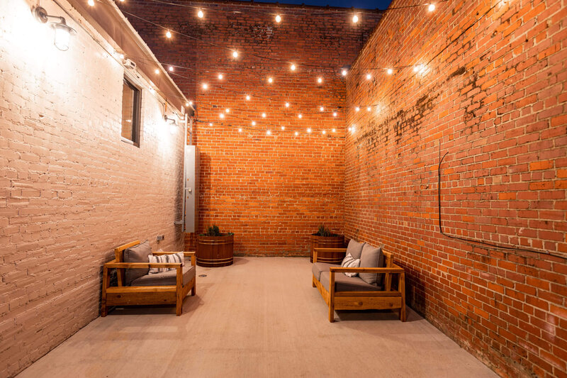 A narrow outdoor patio with string lights overhead, two wooden sofas with cushions on either side, potted plants at the far end, and surrounded by tall brick walls.