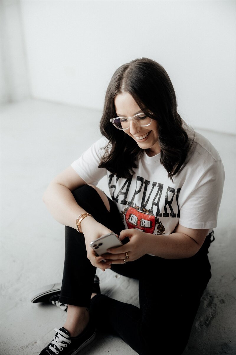 Professional woman seated on a clean white floor, smiling warmly as she engages with her smartphone. Her casual style and relaxed demeanor set the tone for accessible and friendly communication, reflecting the approachable nature of KP Virtual Assistance's client relationships