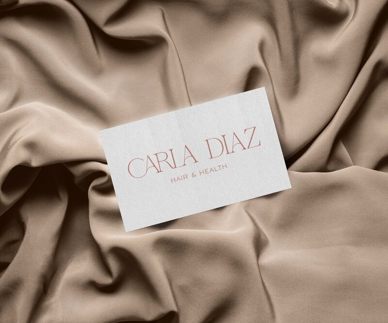 A white card with the text "CARLA DIAZ HAIR & HEALTH" rests on beige, softly draped fabric—an elegant showcase of custom Showit web design and branding.