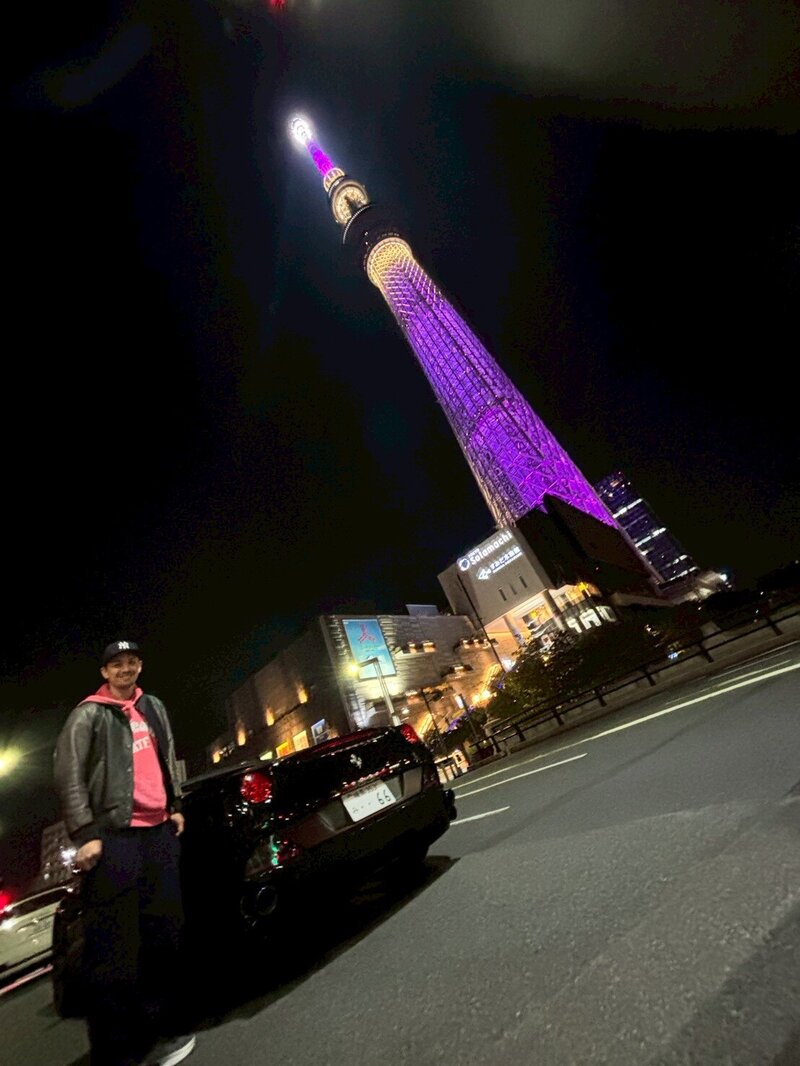 A man standing next to a black Ferrari with Tokyo Skytree illuminated in purple in the background, under the night sky.
