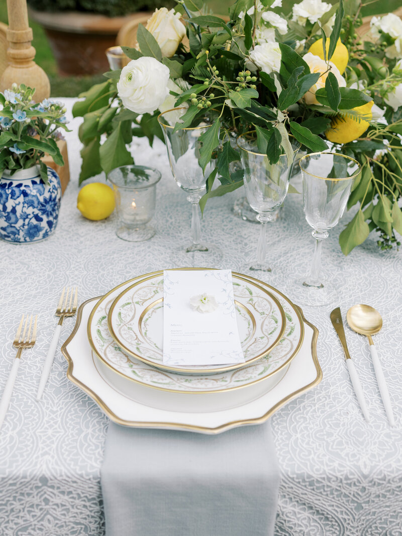 Tablescape details of white and gold place setting with blue chinoiserie vases, green and white florals, and lemons