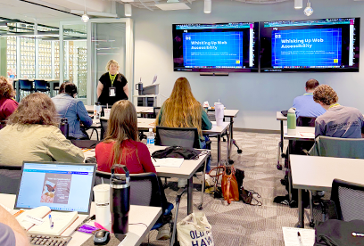 audience POV during a workshop. students are sitting at lecture style tables working on their laptops. 