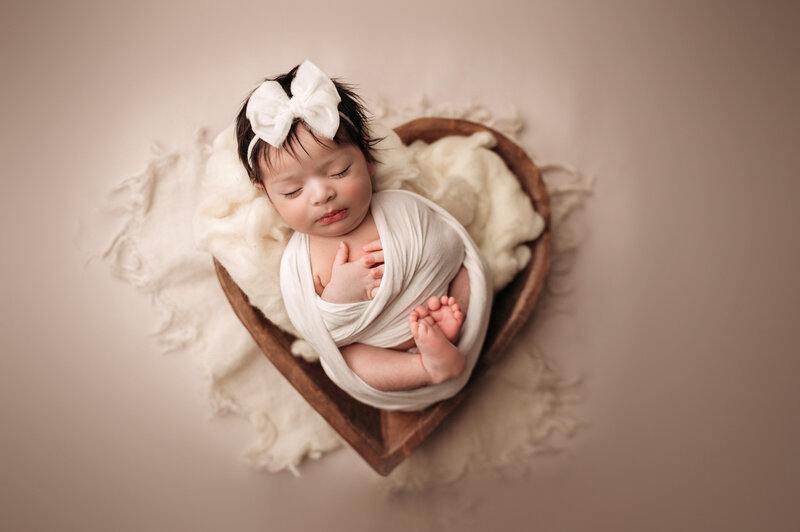 sleeping baby girl in white swaddle sleeping inside of brown heart bowl prop