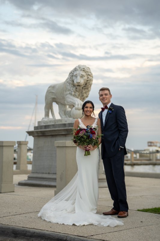 Newlywed couple posing for wedding photos next to ornate stone lion statue at the White Room venue in historic downtown Saint Augustine, with bride in white lace dress holding colorful bouquet and groom in navy suit with red tie, bridge visible in background under cloudy sky