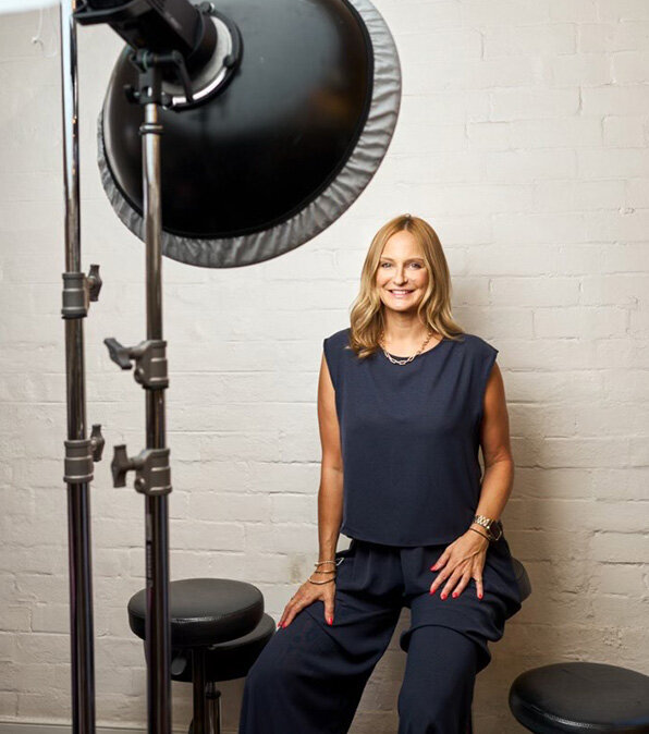 Professional hair educator Katy Reeve smiling in a photography studio, seated next to studio lighting equipment against a white brick wall.