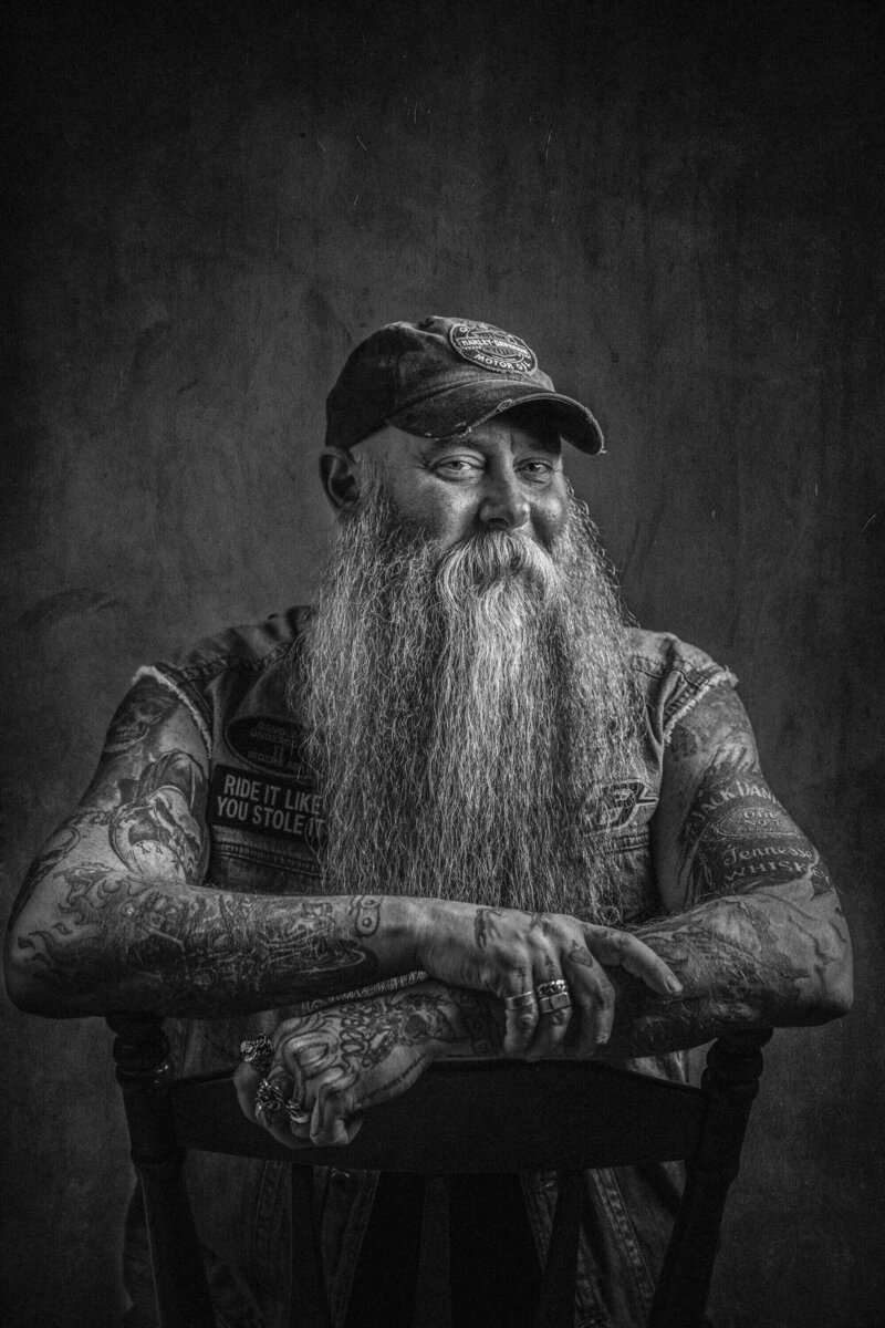 man leans on chair with long beard and tattoos